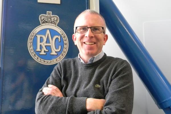 Alistair McMillian standing in front of an RAC logo. He is smiling and has his arms folded.