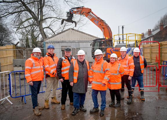 A group of 8 people wearing hi-vis jackets standing in front of a demolition site