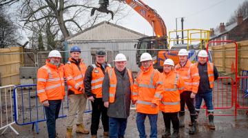 A group of 8 people wearing hi-vis jackets standing in front of a demolition site